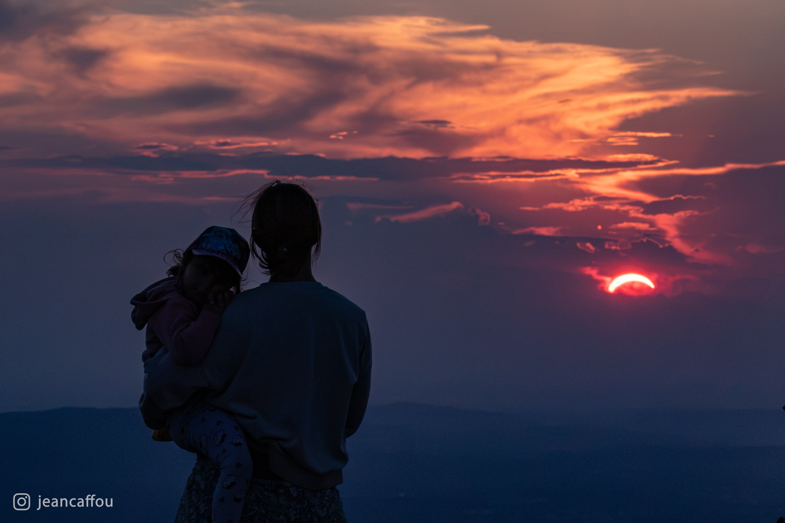 Starš z otrokom v naročju opazuje srpasto Sonce skozi večerno meglico.