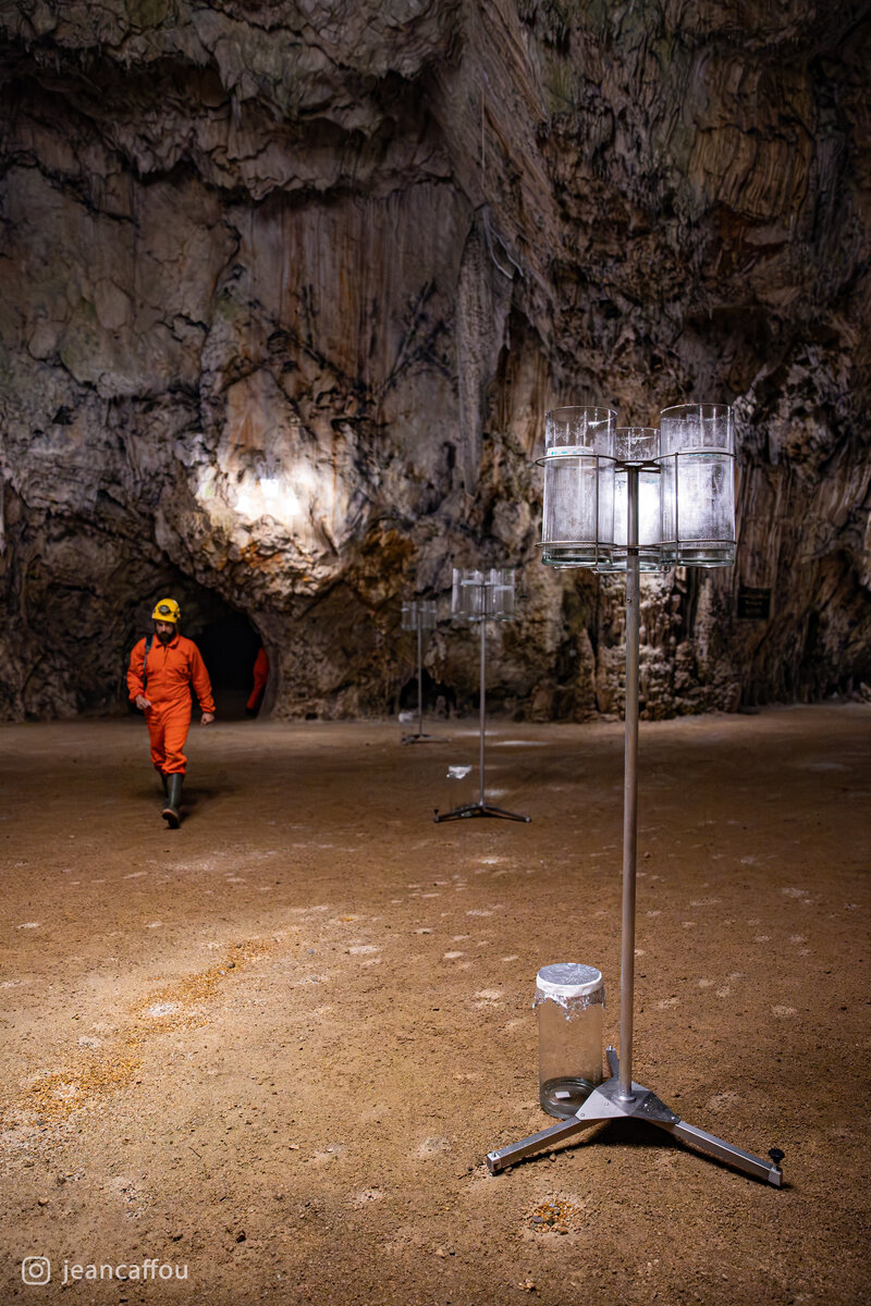 An elevated glass deposition sampler stands beside the path in Postojna Cave, with Blaž Kogovšek approaching in orange coveralls.