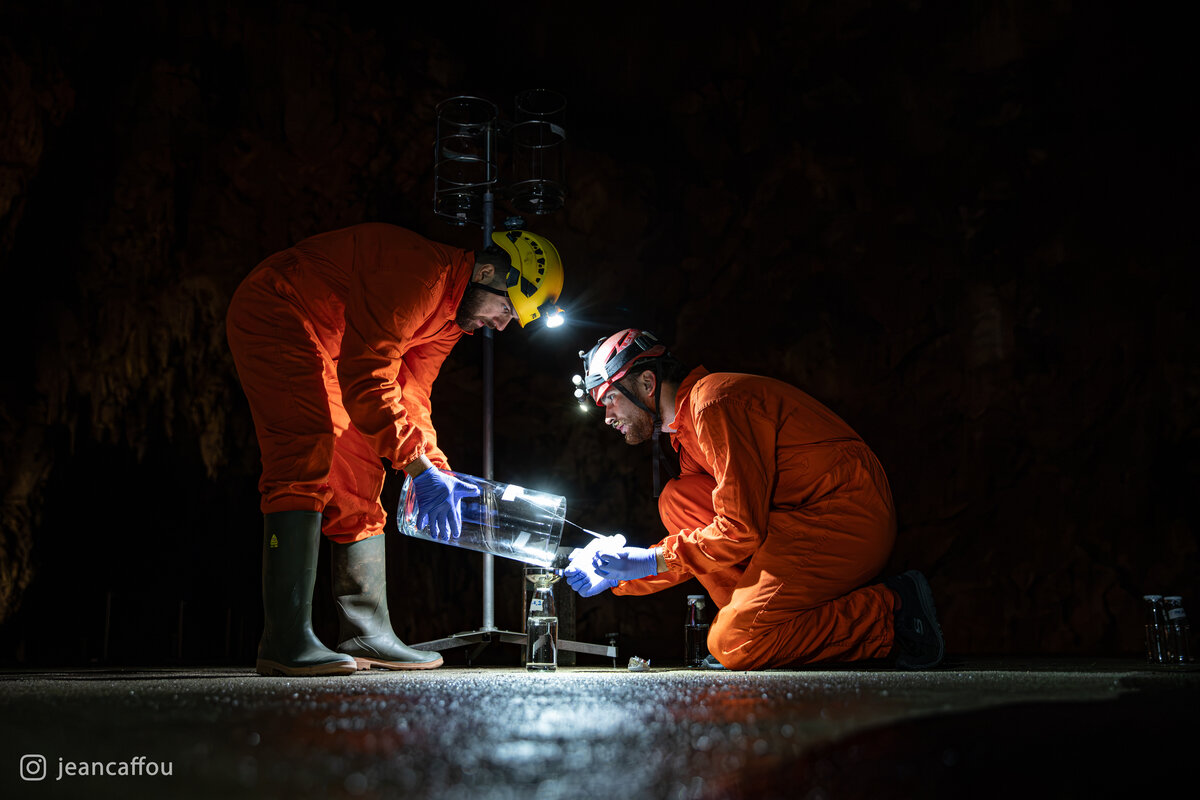 Blaž Kogovšek and Raffaele Bruschi carefully pour a sample between glass vessels, wearing orange cotton coveralls and blue gloves in Koncertna dvorana (Concert Hall) in Postojna Cave.