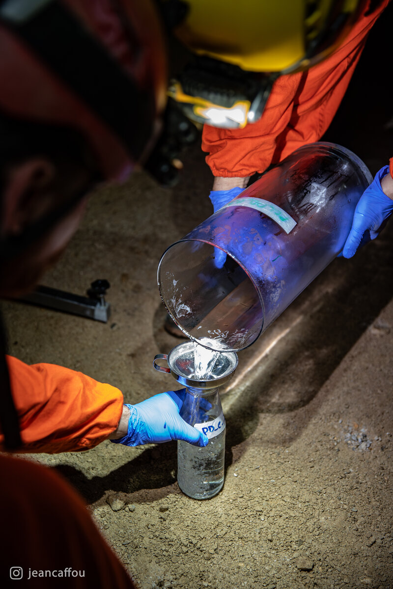 A close view of blue-gloved hands pouring water from a large glass collector into a smaller sample container in Postojna Cave.