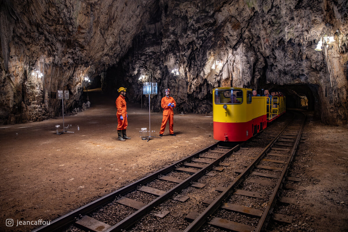 A yellow and red cave train enters Plesna dvorana beside the microplastic sampling station and two researchers in orange coveralls.