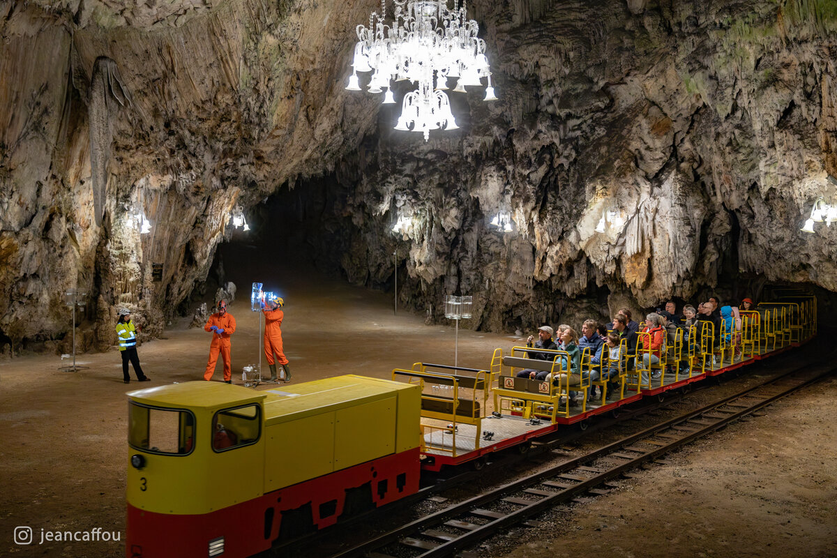 A train filled with visitors crosses Plesna dvorana under its chandelier, with the monitoring station visible beside the railway.