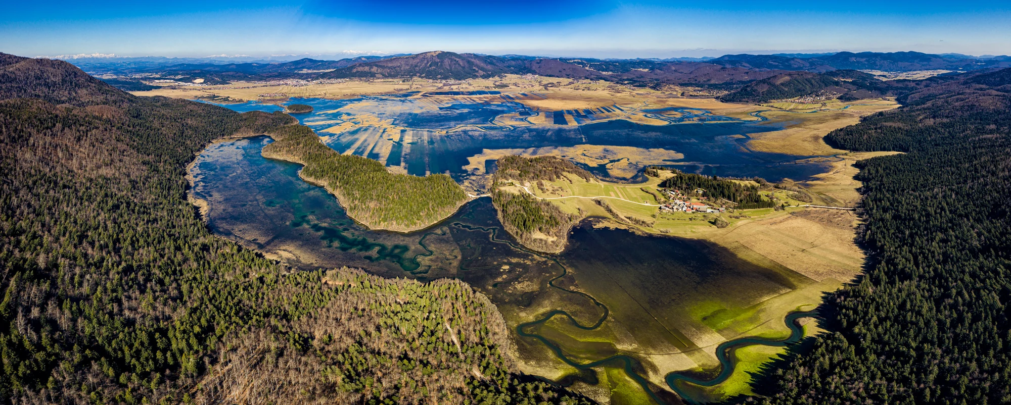 Zračna panorama poplavljenega Cerkniškega polja, kjer vijugaste modre struge obdajajo zelen otok pod oddaljenimi hribi.